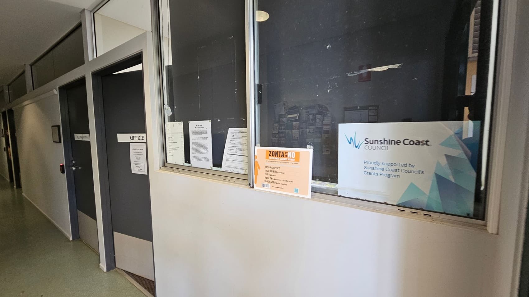A corridor with a green floor and light-coloured walls. On the left, there are two dark doors labelled "OFFICE" and "ENTRY/EXIT". On the right, a window displays several notices, including a "Zonta Says No" poster and a "Sunshine Coast Council" sign, indicating support from the council's grants programme. The ceiling is not visible.
