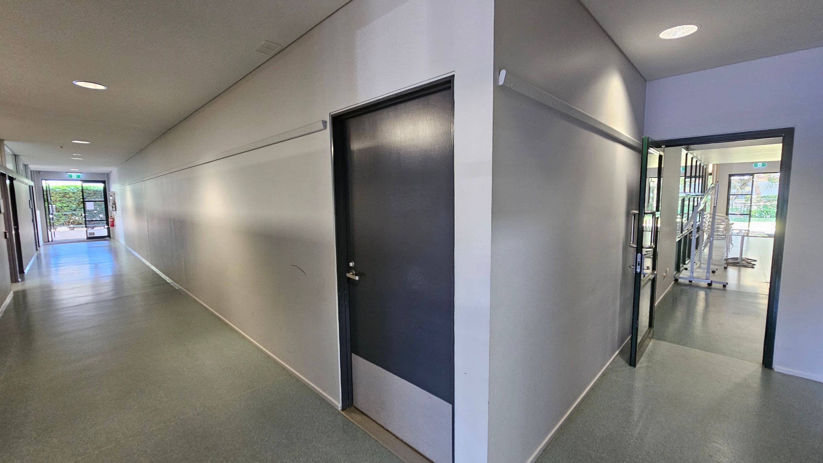 A corridor with grey flooring and white walls. On the left, a closed dark door is set into the wall. The corridor extends towards a glass door at the end, leading outside with greenery visible. On the right, an open door leads into a room with shelves and stacked chairs. The ceiling has recessed lighting.