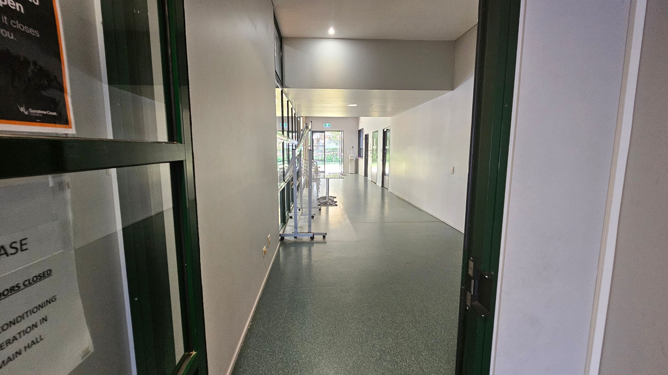 A hallway with a speckled grey floor and white walls. On the left, there's a glass door with a sign. Further down the left side, a row of windows and a metal rack with stacked chairs. On the right, several closed doors line the wall. At the end of the hallway, a glass door leads outside, with visible greenery through it. The ceiling is plain with recessed lighting.