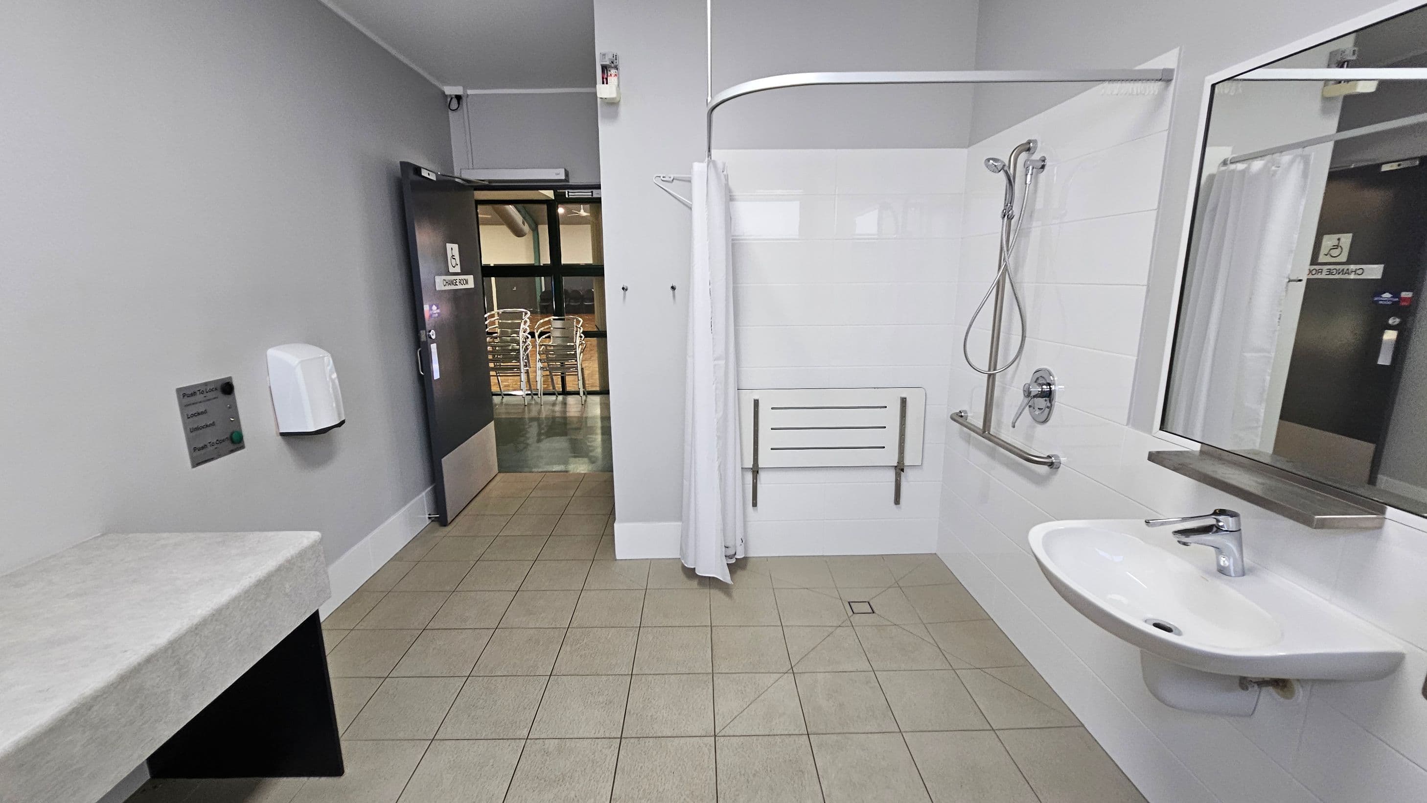 A spacious, accessible bathroom with a light green tiled floor. On the right, there's a white sink with a mirror above it and a shower area with a handheld showerhead, grab bar, and a foldable seat. A white curtain partially encloses the shower. On the left, a grey wall features a soap dispenser and a control panel. The door is open, revealing a hallway with stacked chairs visible outside. The ceiling is plain and white.
