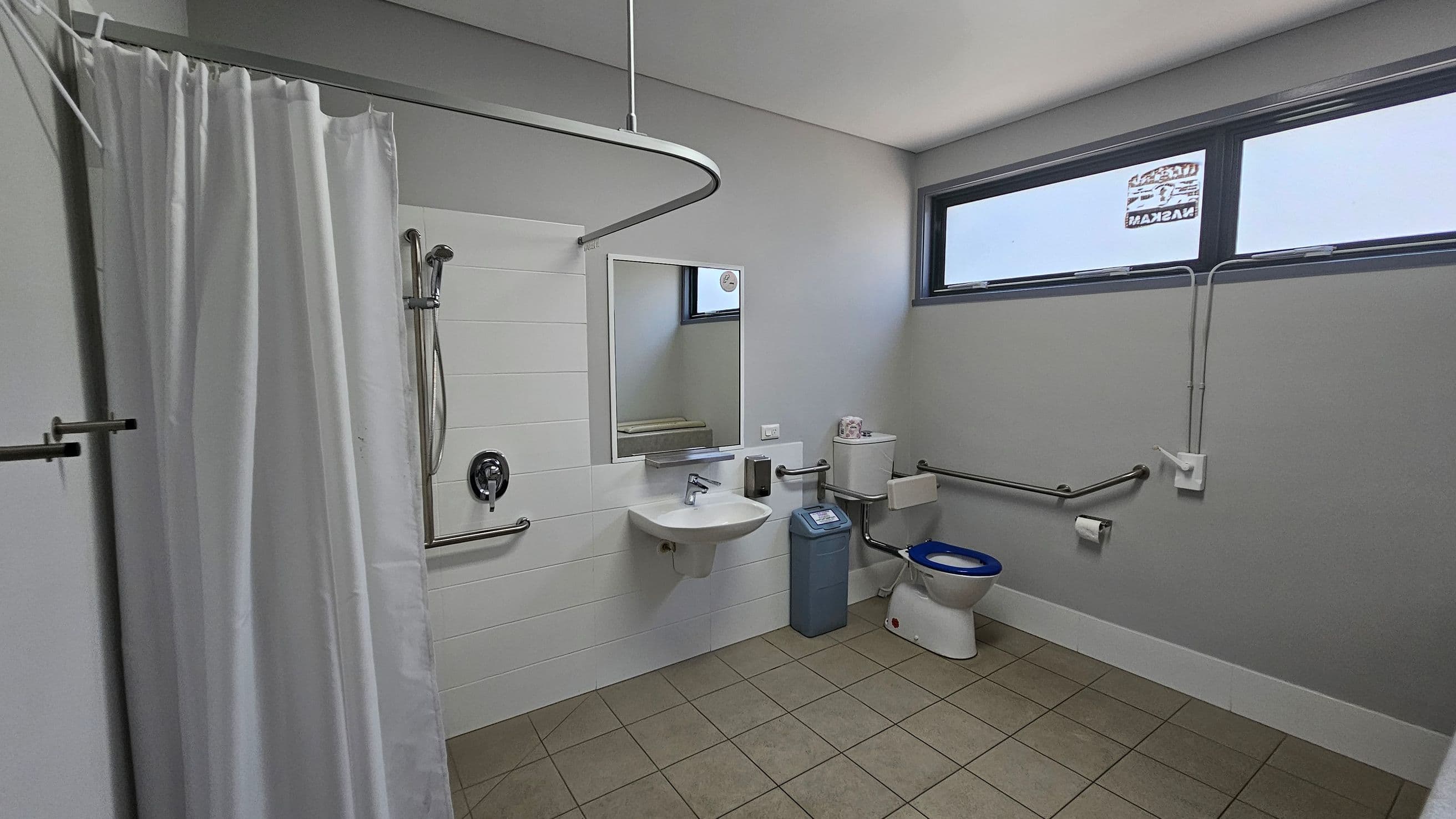 A bathroom with a green tiled floor and a white ceiling. On the left, there's a shower area with a white curtain and a wall-mounted showerhead. In the centre, a wall-mounted sink with a mirror above it is flanked by a soap dispenser. To the right, a toilet with a blue seat is next to a blue bin. The room has grab bars for accessibility and a high horizontal window on the right wall.
