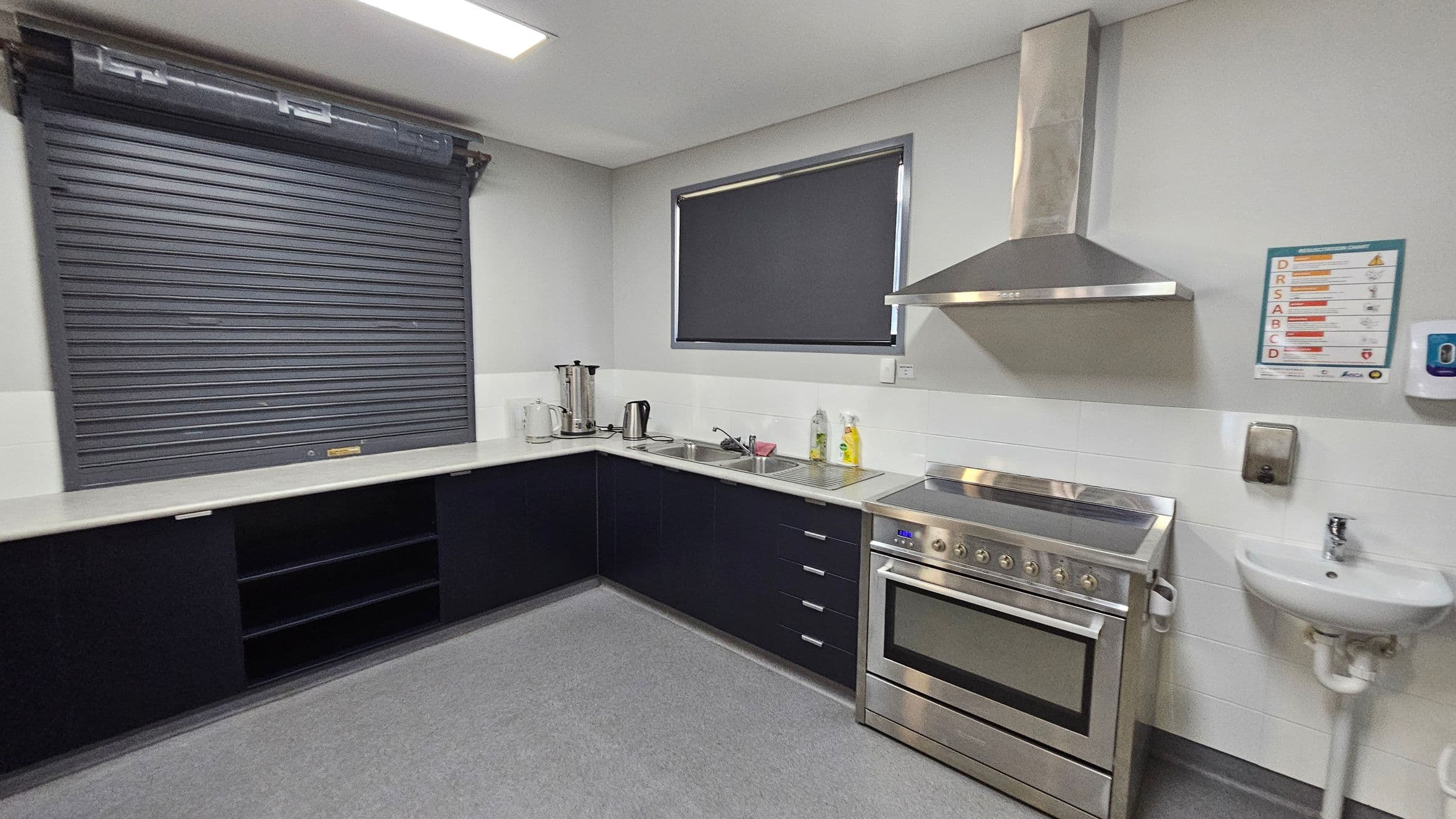 A modern kitchen with a grey roller shutter on the left above black cabinets. In the middle, there's a countertop with a kettle, a coffee maker, and a sink. On the right, a stainless steel oven with an overhead extractor fan is next to a small white sink. A window with a dark blind is above the counter. The floor is grey, and the ceiling has a rectangular light fixture. A safety instruction poster and a soap dispenser are mounted on the wall.