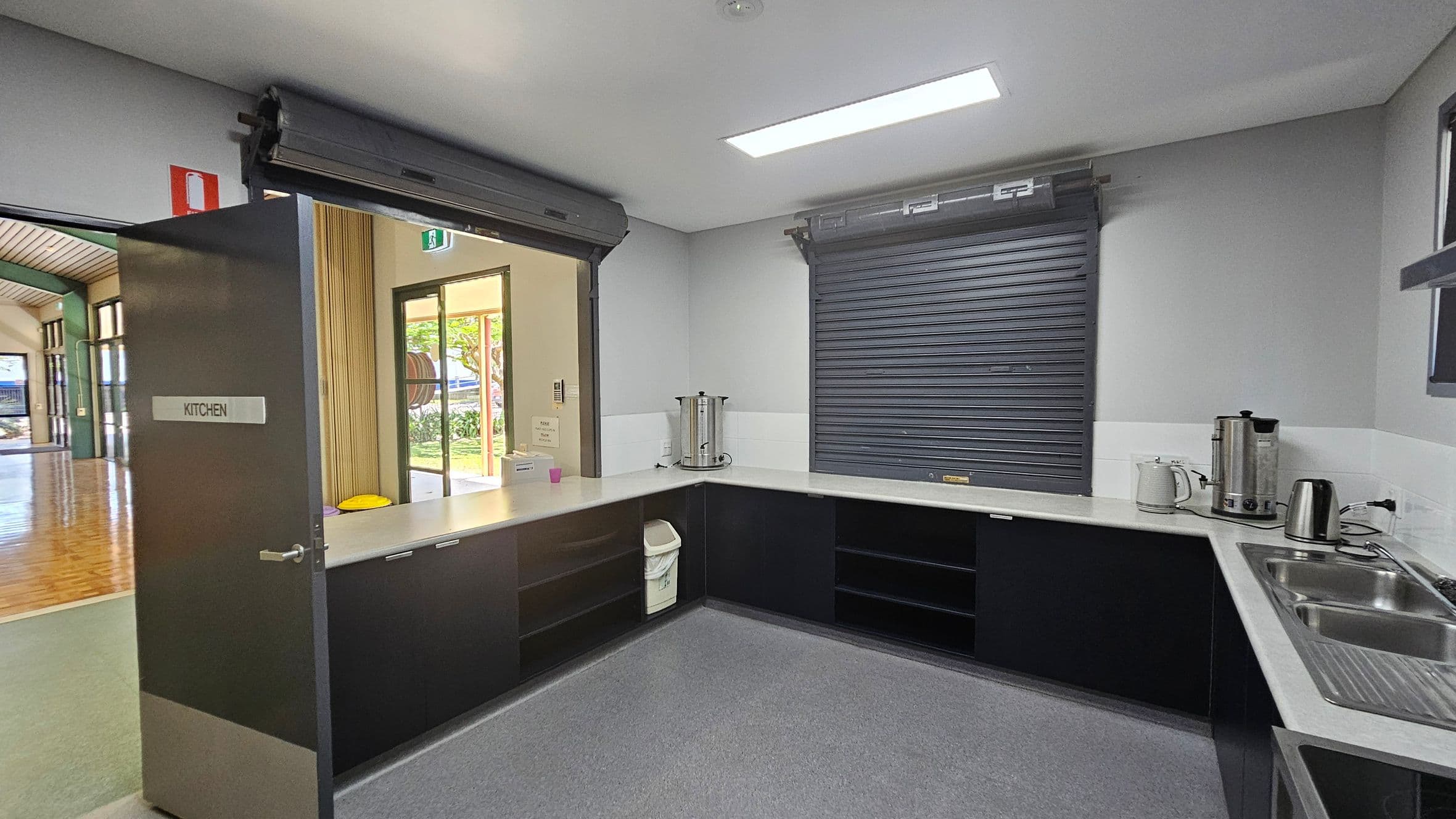 A kitchen with a grey door on the left labelled "KITCHEN" leading to a hallway with wooden flooring. The kitchen has a U-shaped counter with a stainless steel sink on the right. Two large metal urns, a kettle, and a toaster are on the counter. A roller shutter window is closed above the counter. The floor is grey, and the ceiling has a rectangular fluorescent light. A bin is tucked under the counter.