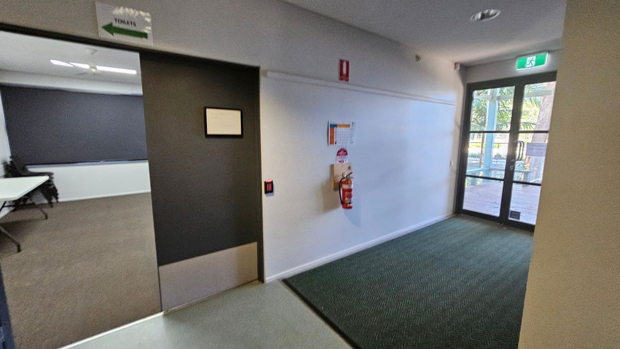 A hallway with a grey and green floor. On the left, there is a glass door with an exit sign above it and a row of grey lockers. The right side features a long white wall with windows near the ceiling, and several doors leading to rooms. The ceiling is white with recessed lighting. At the far end, another glass door is visible, leading outside.