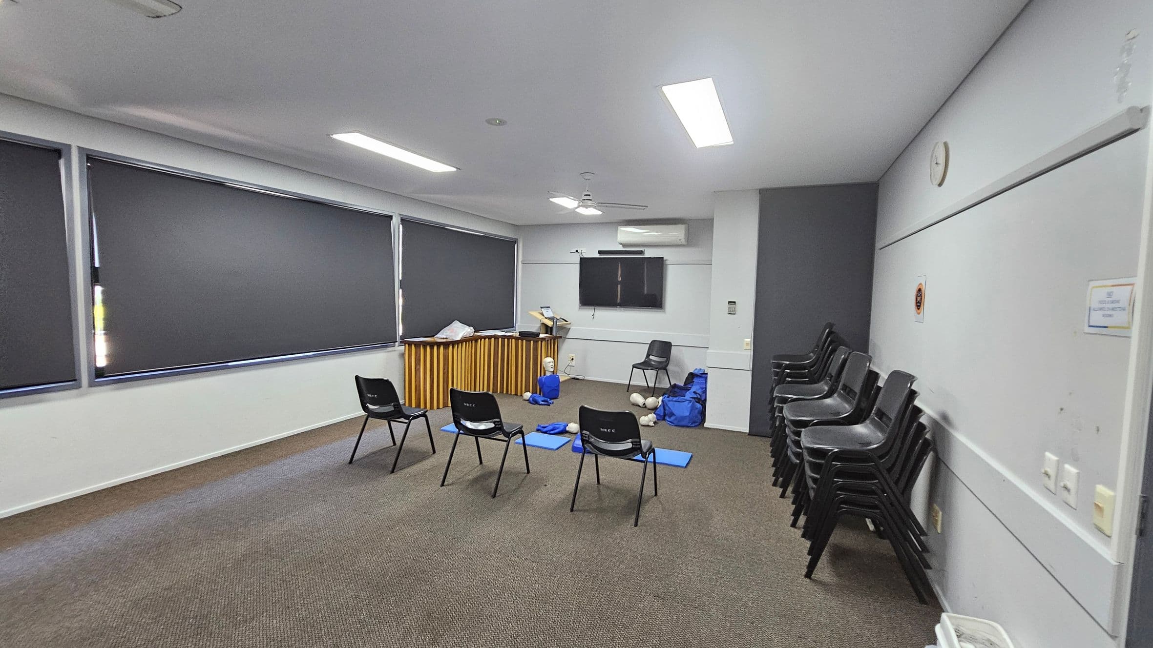 A room with grey walls and a patterned carpet floor. On the left, there are large windows with dark blinds. In the centre, several black chairs are arranged in a semi-circle, with blue mats and CPR mannequins on the floor. At the back, a wooden counter holds various items, and a wall-mounted TV is above it. On the right, a stack of chairs is against the wall, and a clock is visible. The ceiling has fluorescent lights and a fan.