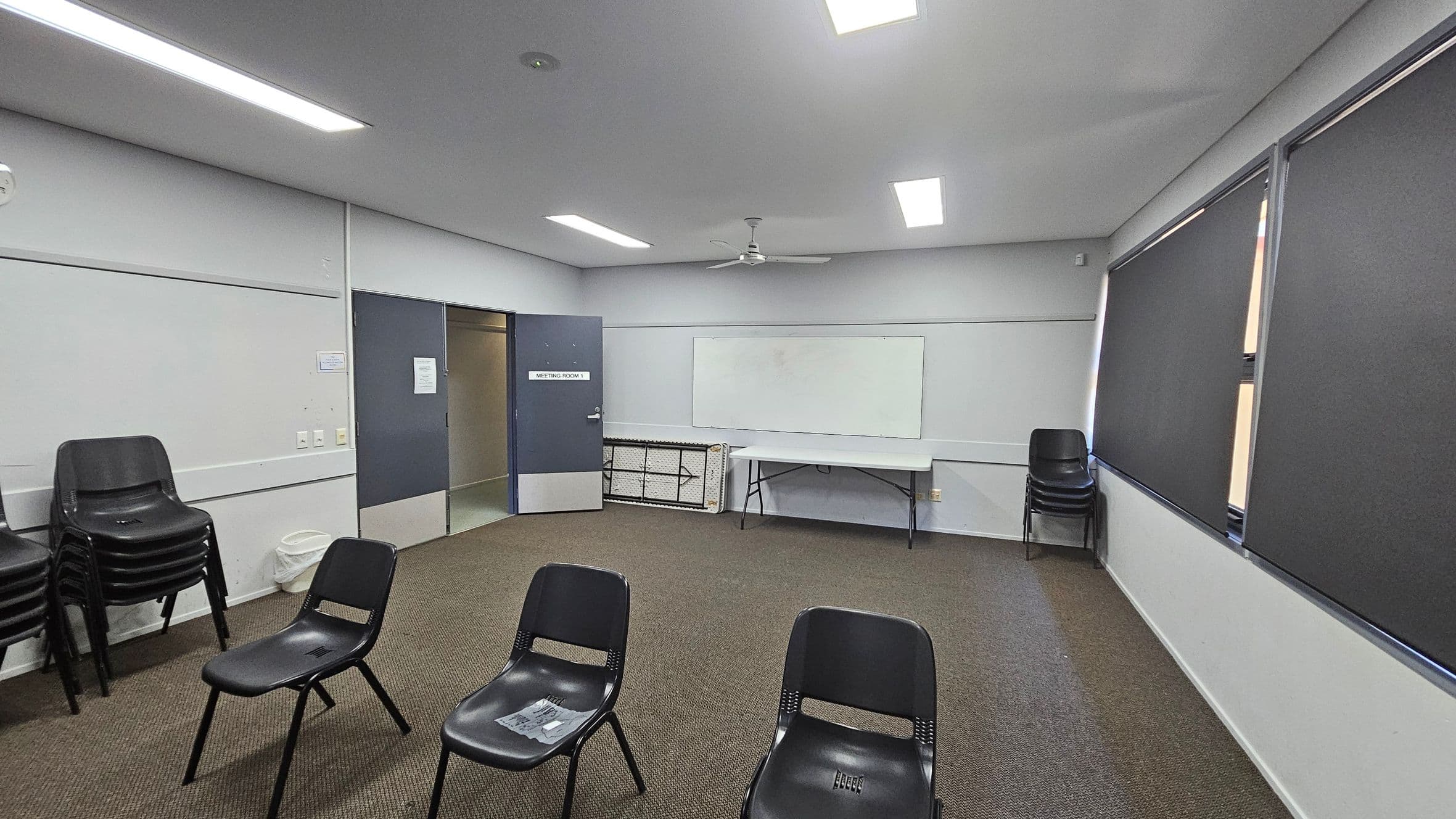 A meeting room with a grey ceiling and fluorescent lights. The floor is carpeted in a mix of green and brown. On the left, there is a stack of black chairs and a waste bin. In the centre, several black chairs are arranged in a semi-circle. The back wall has a whiteboard and a folding table. The right side features windows with dark blinds. A door labelled "Meeting Room 1" is open on the left wall.