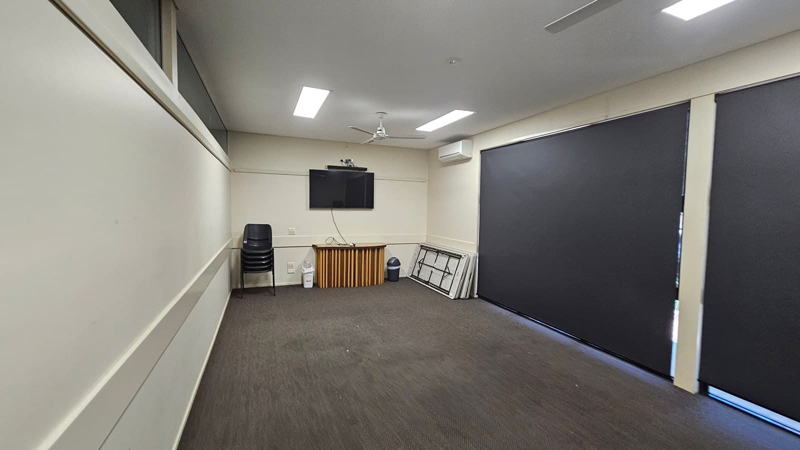 A meeting room with a brown carpeted floor and a white ceiling featuring two ceiling fans. On the right, a door labelled "MEETING ROOM 2" is partially open. Inside, a wooden table is visible. Against the far wall, there are large black blinds covering the windows. To the left, a stack of folded tables and a bin are placed in the corner. A stack of black chairs is positioned near the centre of the room.