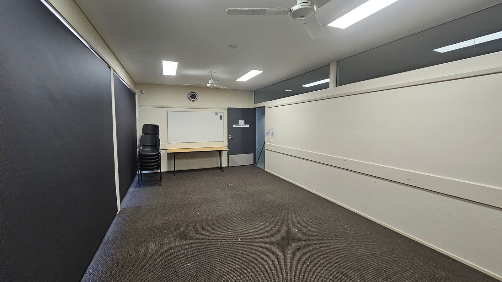 A small room with a dark carpeted floor and light green walls. On the left, there are large black panels. At the back, a stack of black chairs is next to a wooden table beneath a whiteboard. Above the whiteboard, a clock is mounted on the wall. A grey door labelled "Meeting Room 2" is to the right of the whiteboard. The ceiling has fluorescent lights and two ceiling fans. The right wall has a high window strip.