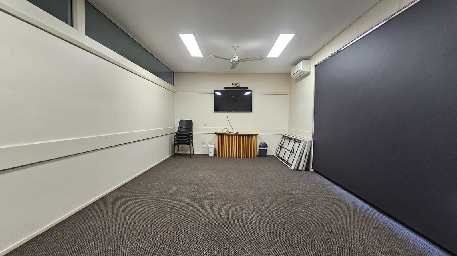 A room with light-coloured walls and a dark carpeted floor. On the far wall, a flat-screen TV is mounted above a small wooden cabinet. To the left, a stack of black chairs is placed against the wall. On the right, several folded tables are leaning against the wall next to a bin. The ceiling has two rectangular lights and a ceiling fan. An air conditioning unit is mounted on the right wall.
