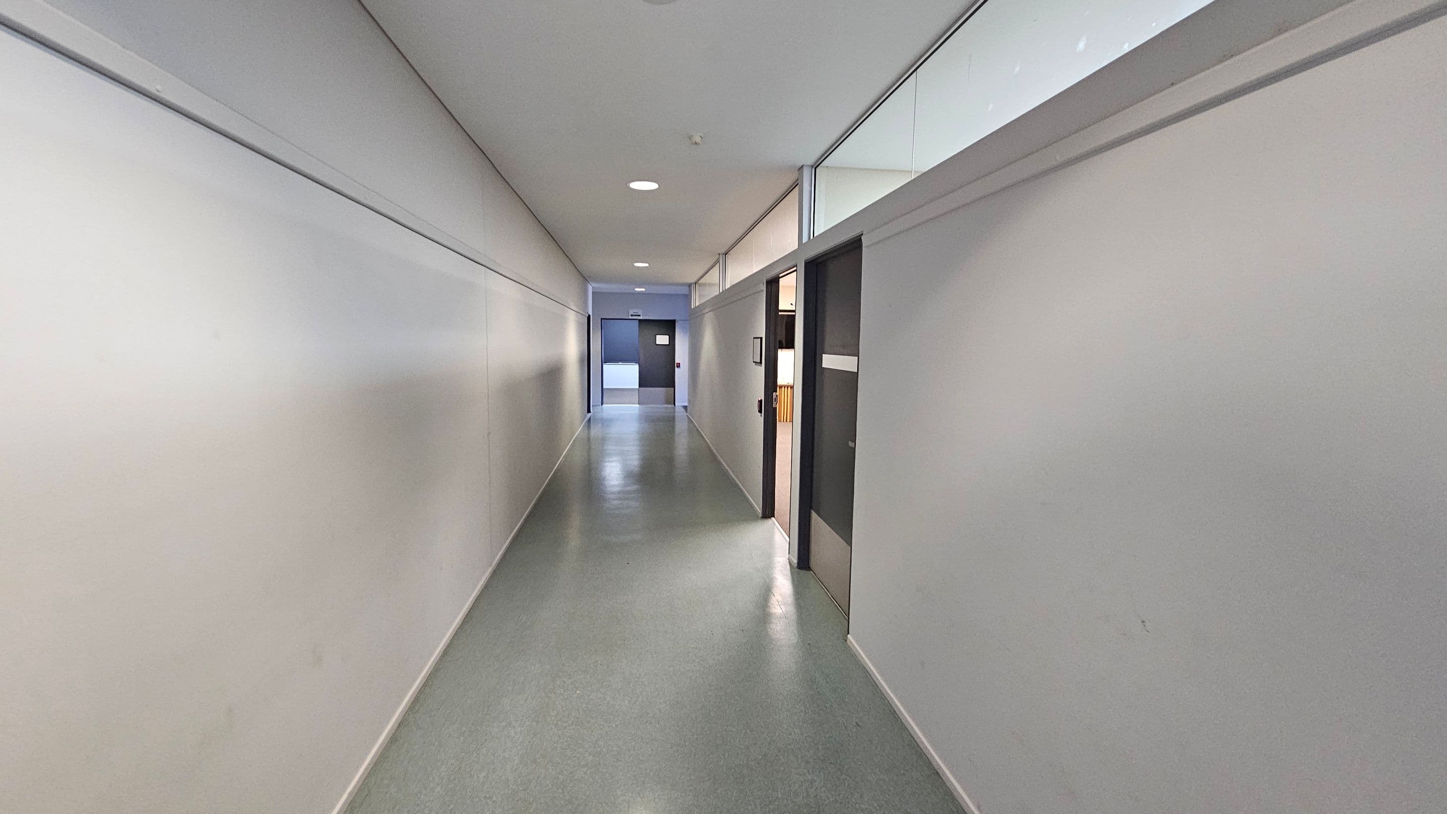 A long, narrow corridor with smooth, light grey walls and a polished grey floor. On the right, there are several doors, one of which is open, revealing part of an interior space. Above the doors, there are glass panels. The ceiling is white with recessed lighting. At the end of the corridor, a closed door is visible.