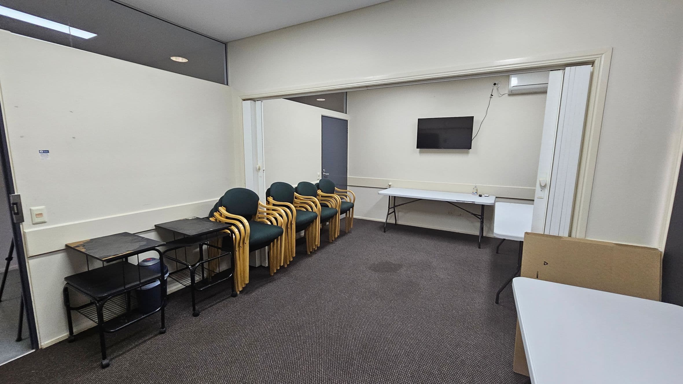 A room with a grey carpeted floor and white walls. On the left, there are three black metal tables with shelves, and a stack of wooden chairs with green cushions. In the centre, a doorway leads to another area with a wall-mounted TV and a long folding table. On the right, a white folding table is partially visible. The ceiling is plain white, and there is a small air conditioning unit above the TV.