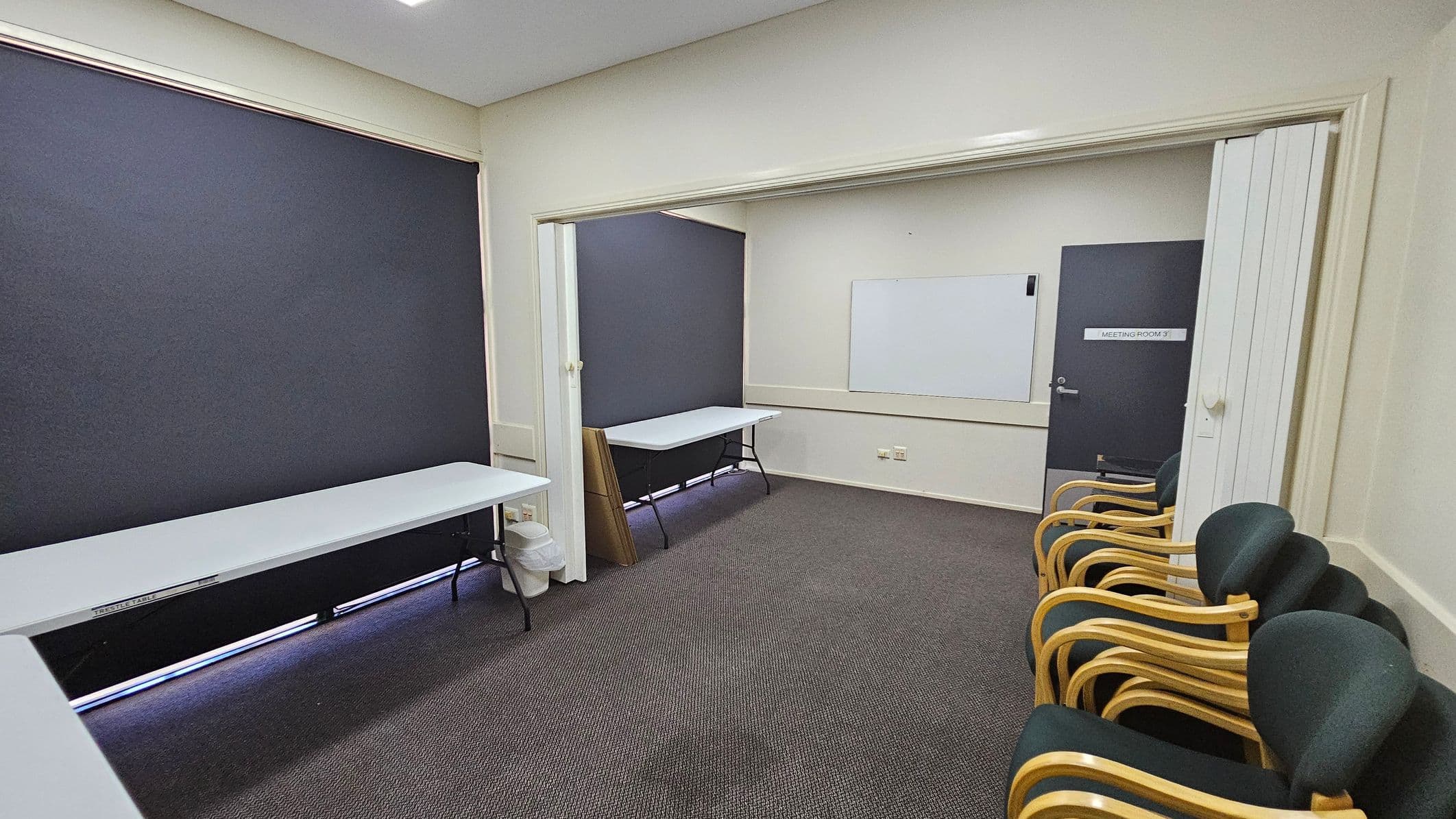 A room with grey carpet and light-coloured walls. On the left, there is a long white table against a wall with a dark blind. In the middle, a partition separates two areas, each with a similar table. On the right, a row of chairs with wooden frames and dark upholstery is lined up against the wall. A door marked "Meeting Room 3" is visible, along with a whiteboard on the wall. The ceiling is plain with recessed lighting.