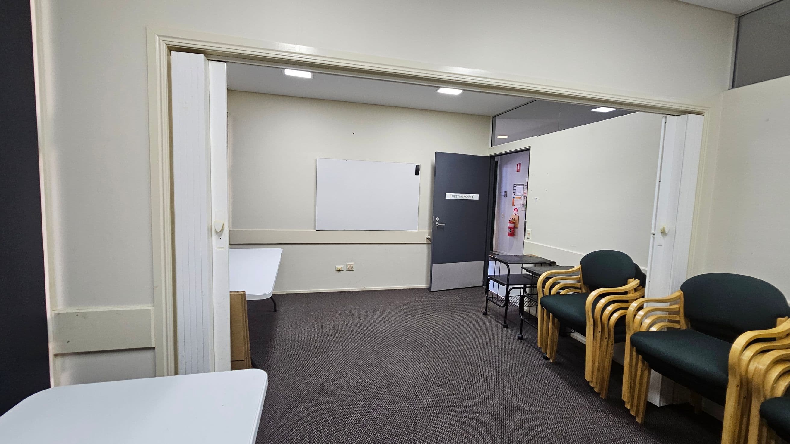 A room with grey carpet and white walls. On the left, there are white tables. In the centre, a whiteboard is mounted on the wall, and a door with a sign leads to another room. On the right, there are stacked wooden chairs with dark green cushions and a small black metal table. The ceiling has recessed lighting.