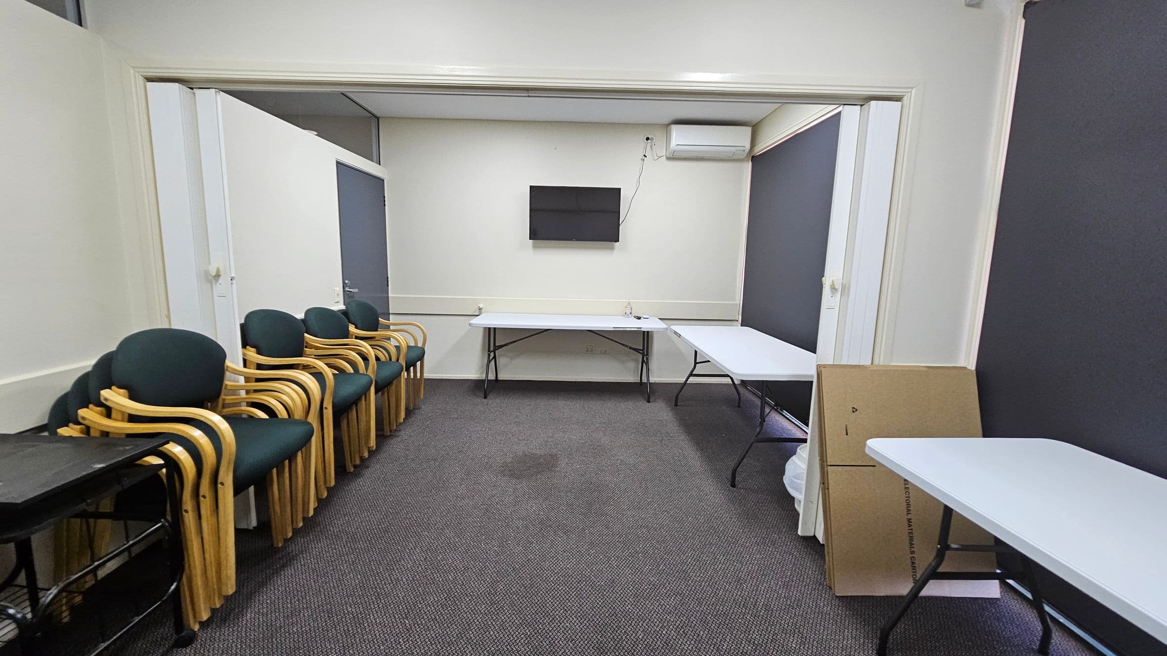 A room with a grey carpeted floor and white walls. On the left, there is a stack of wooden chairs with green cushions next to a black trolley. In the centre, a wall-mounted television is above two white folding tables. On the right, there are more tables and a cardboard box leaning against the wall. An air conditioning unit is mounted on the wall above the tables. The ceiling is plain white.