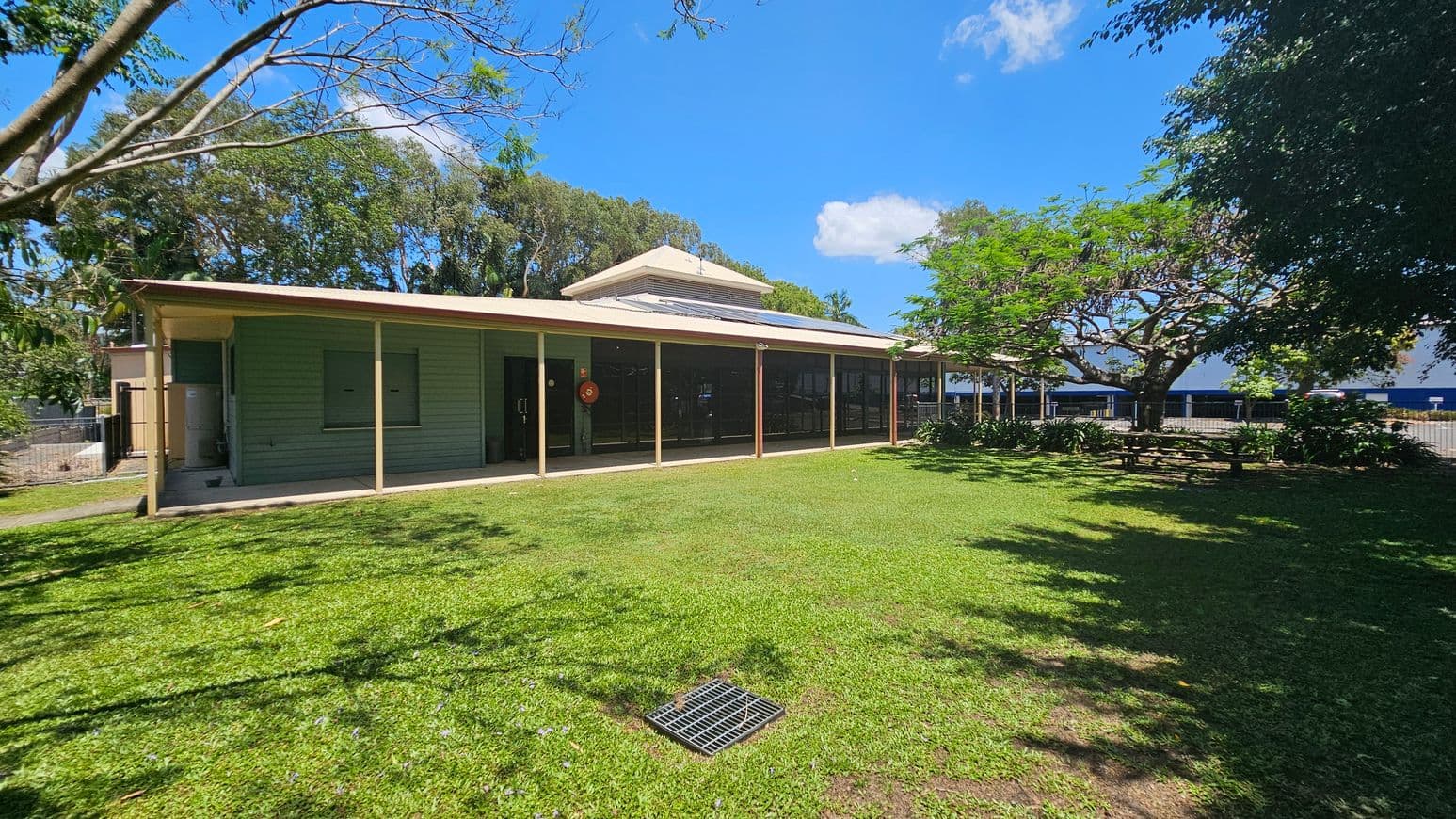 A single-storey building with a green exterior and a beige roof is situated in the centre. It has a covered veranda with several support posts. To the right, there is a large tree providing shade over a grassy area. In the foreground, a well-maintained lawn with a metal grate is visible. The background features more trees and a glimpse of a blue structure. Shadows from the trees are cast on the grass.
