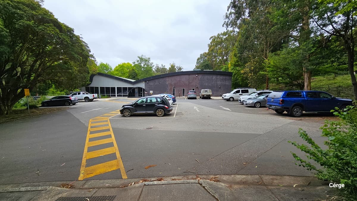 Parking spaces in Monbulk Aquatic Centre nestled in a wooden area. The park space floor transitions from dark grey to light grey and back to dark grey when going closer to the entrance. On the floor surface of the parking area are white directional arrows and yellow lines marking the parking spaces. Monbulk Aquatic Center has a curved roof with grey to dark purple brick wall finish. The entrance can be distinguished by a black arch near the glass windows with white metal frames.