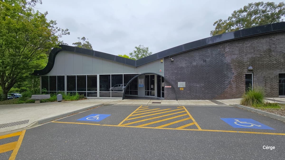 Two accessible car parking spaces in front of Monbulk Aquatic Centre are marked by a blue background with a white wheelchair image. Between the 2 accessible car park spaces are yellow diagonal lines enclosed by a yellow rectangle. The building has a large glass window on the left side and a smaller window on the right side. The entrance is in the center of the building and has a black arch above it.