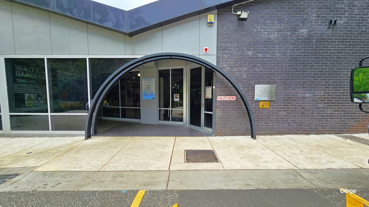 Entrance with two black metal arches at Monbulk Aquatic Centre. The entrance is a curved archway with a glass door and windows. The building transitions from grey tile with glass windows that have white metal frames to dark purple brick from left to right. The pathway in front is made of concrete tiles and is connected to the grey tiles of the entrance floor. A white sign on the brick wall has red HAZCHEM detailed on it.