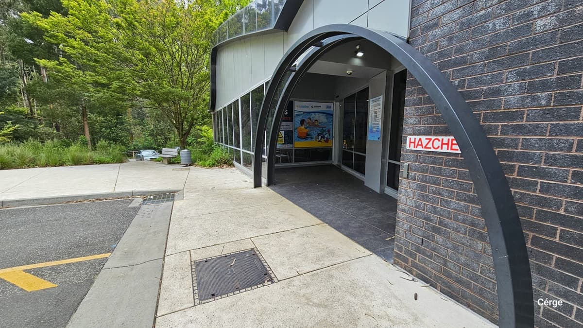 The side view of the entrance with two metal curved archways. The entrance is flanked by two large windows, one of which has a colorful mural on it. The entrance is also marked by a red and white “HAZCHEM” sign. The building is surrounded by lush greenery and a concrete walkway. Under the tall tree with a large tree canopy cover is a grey metal bench, and beside it is a grey circular perforated garbage bin.