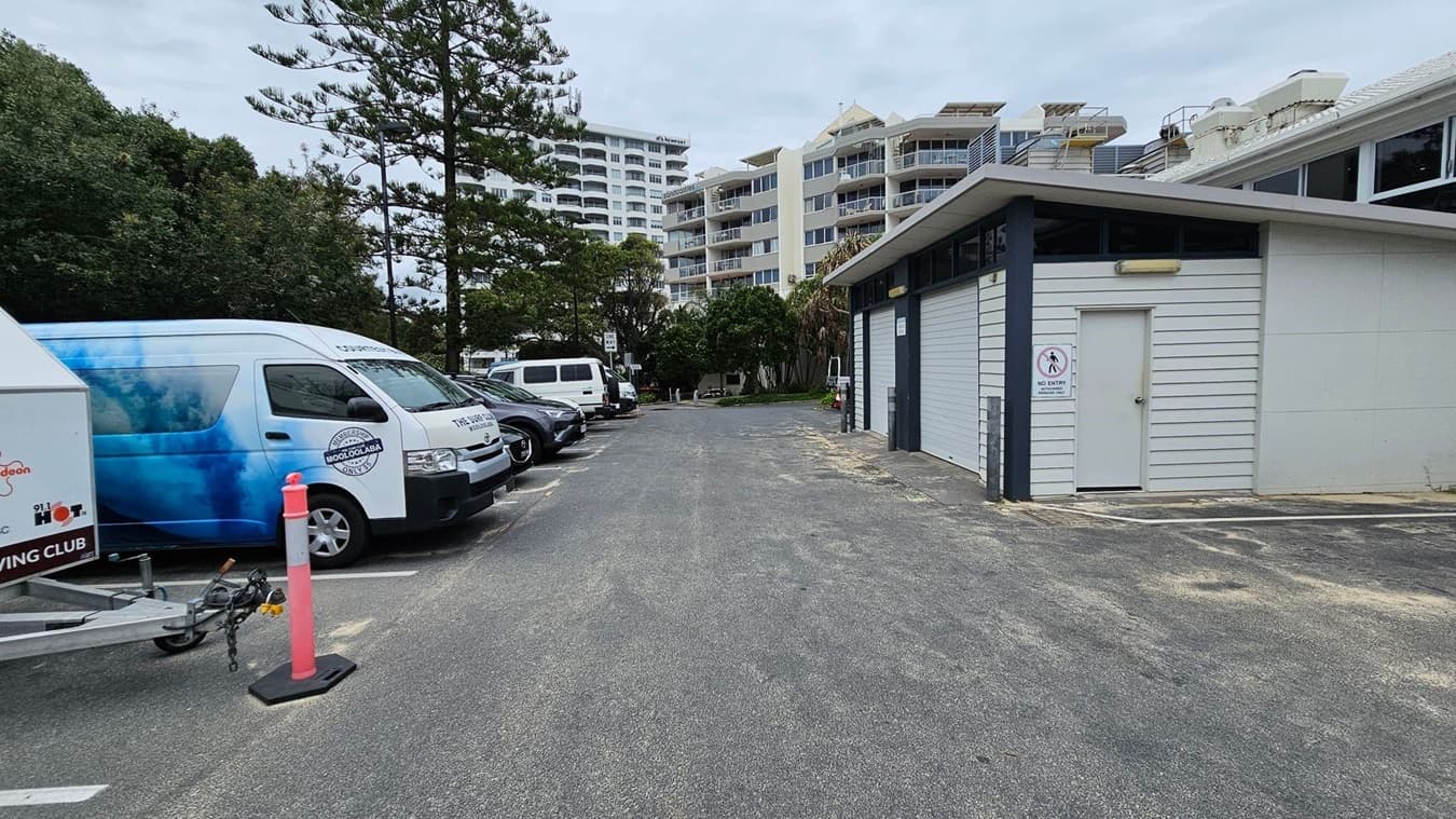 A car park with several vehicles on the left, including a van with a blue design. A trailer is attached to one vehicle, and an orange traffic cone is nearby. On the right, there's a building with a white exterior and a "No Entry" sign on the door. In the background, there are tall trees and multi-storey residential buildings. The ground is paved, with some patches of green moss or grass.