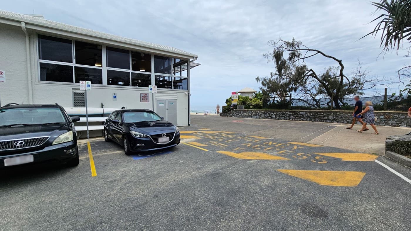 A parking area with two cars, a Lexus and a Mazda, parked in front of a building with large windows on the left. The ground has yellow markings indicating a tow-away zone. On the right, a stone wall lines a path where two people are walking. In the background, there are sparse trees and a glimpse of the beach and ocean.