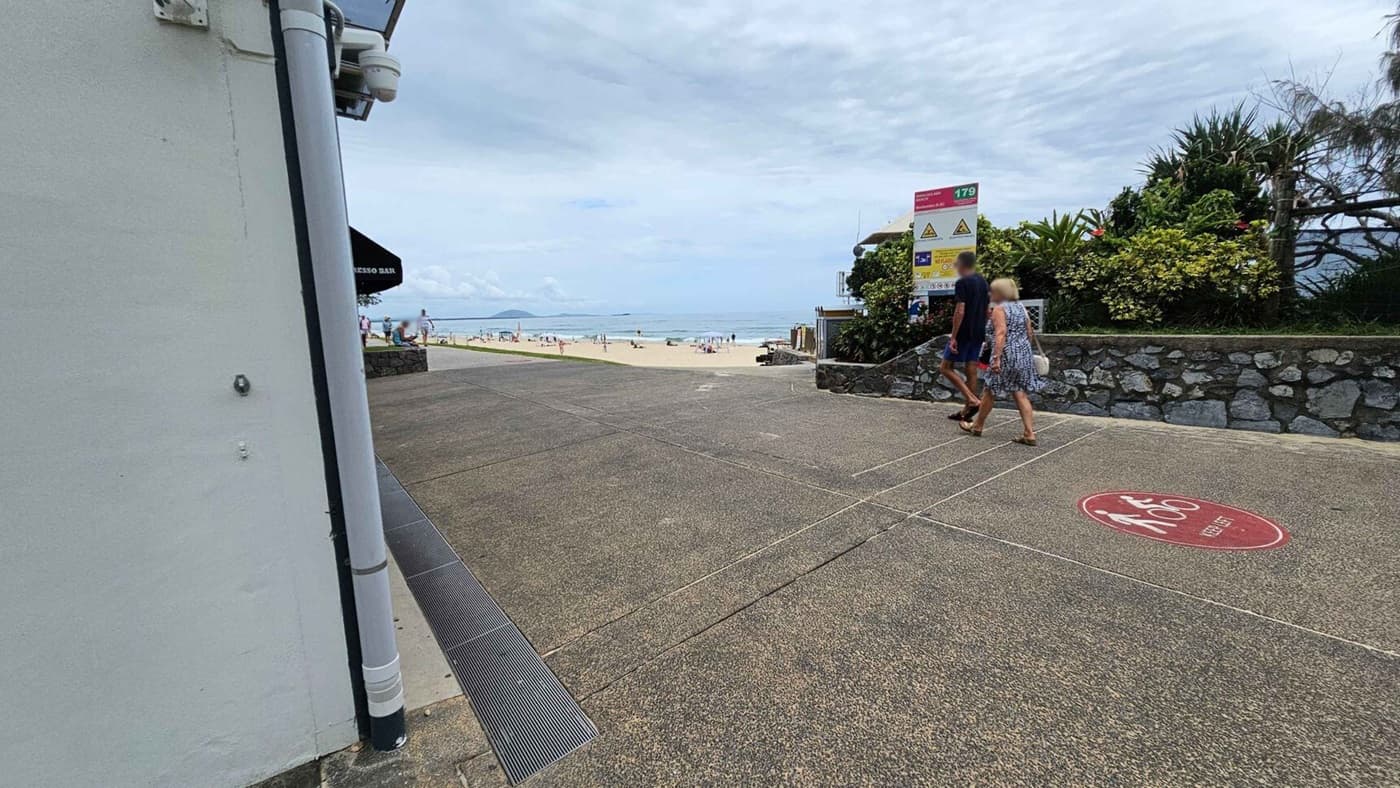 A beach scene with a pathway leading to the sand. On the left, there's a building with a pipe and a sign for an espresso bar. On the right, a stone wall with lush greenery and a sign with safety information. Two people walk along the path, which has a textured surface and a red circle with "Keep Left" written on it. In the background, people are on the beach near the ocean.
