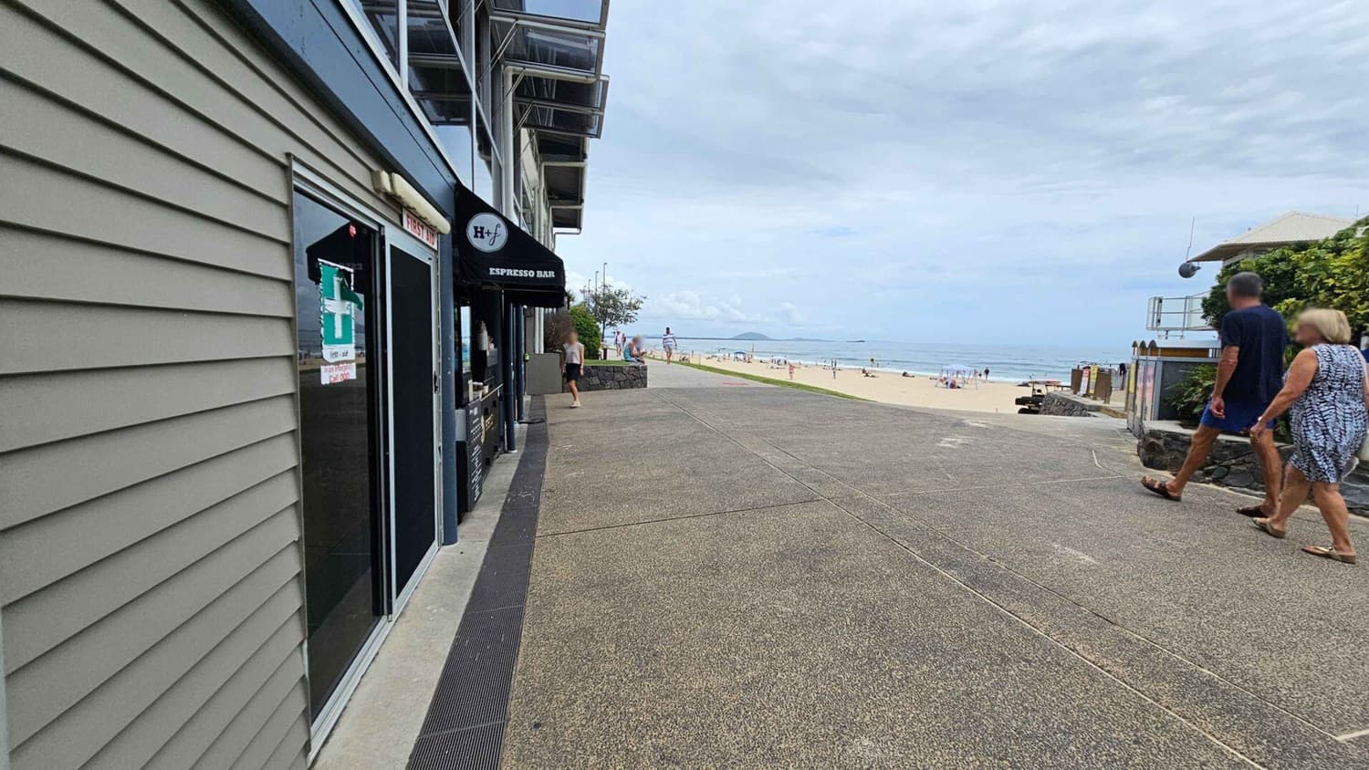 A beachside scene with a grey building on the left featuring a sign for an espresso bar. The pavement is a mix of green and brown tones. In the middle distance, a person walks towards the beach, which is visible on the right with people enjoying the sand and sea. Two people walk along the path on the right, with a stone wall and greenery nearby. The ocean stretches out in the background.