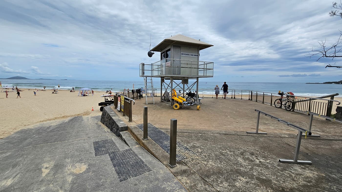 A beach scene with a lifeguard tower on the right, elevated on metal stilts with a ladder. Below it, a yellow rescue vehicle is parked. A few people are walking and relaxing on the sandy beach to the left. The ocean is in the background, with gentle waves. A concrete path with metal railings leads to the beach, and a bicycle is parked near the railings. The sky is overcast.