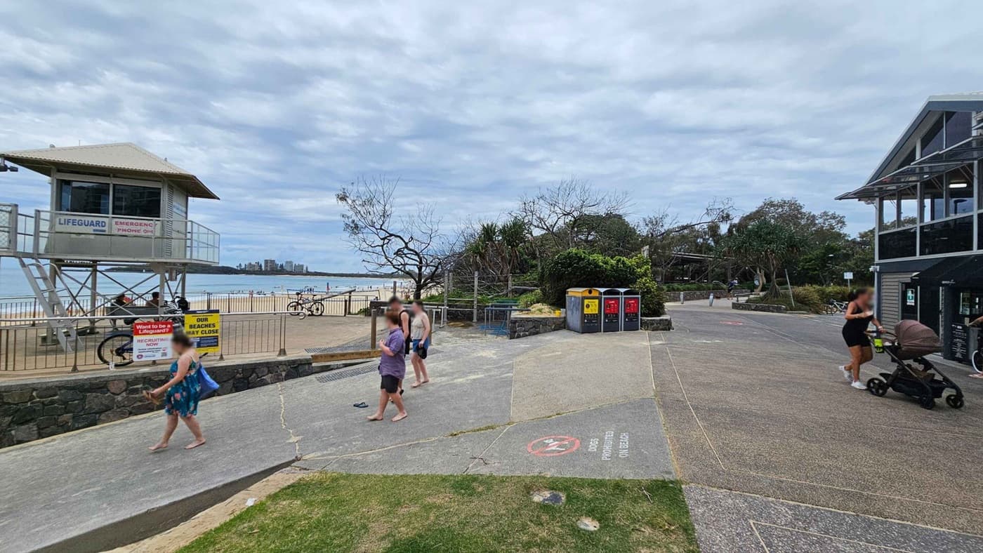 On the left, a lifeguard tower overlooks a sandy beach with a few people and bicycles nearby. Signs about lifeguard recruitment and beach closures are visible. In the centre, a path with people walking leads to the beach. On the right, a woman pushes a pram near a building with a sloped roof. Three bins are positioned along the path, and there are trees and bushes in the background. The ground is a mix of concrete and grass.