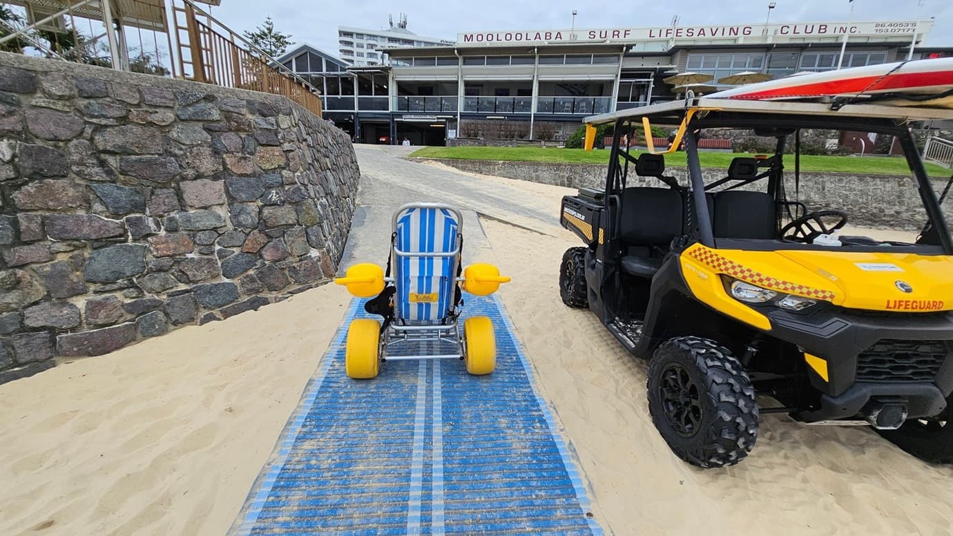 A sandy beach with a blue textured mat occupies the foreground. On the left, a curved stone retaining wall slopes down towards the mat. In the middle, a striped blue and white beach wheelchair with yellow wheels is centred on the mat. On the right, a yellow lifeguard utility vehicle with rugged tyres is parked beside the mat. In the background, a grey stone path leads uphill to the Mooloolaba Surf Lifesaving Club building, with grassy areas and railings on either side.
