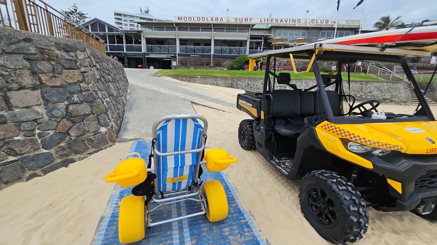 A blue textured mat lies on the sandy floor in the foreground, with a striped beach wheelchair featuring yellow wheels in the centre. To the right, a yellow lifeguard utility vehicle with rugged black tyres rests on the sand. On the left, a stone retaining wall curves upwards next to a sloping concrete ramp. In the background, the Mooloolaba Surf Lifesaving Club building sits on a grassy area with railings, stairs, and pathways leading up to it.