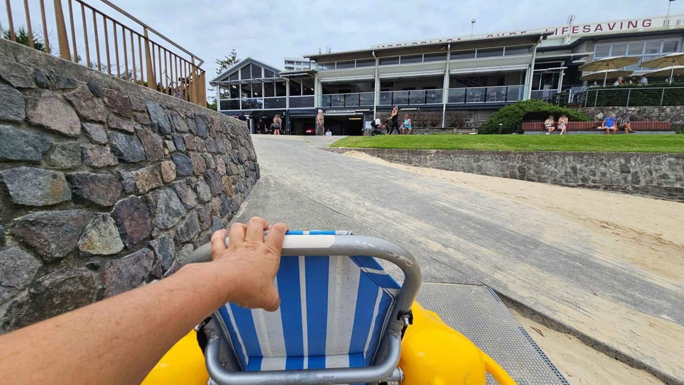 A textured stone wall lines the left side of a sloping concrete pathway. On the right, a sandy section borders a low stone retaining wall, leading to a grass area where people are sitting on benches. In the middle, a hand grips the back of a striped blue-and-white wheelchair with yellow wheels. In the background, a multi-windowed building with a large terrace has several people gathered near its entrance, along with small landscaped bushes.