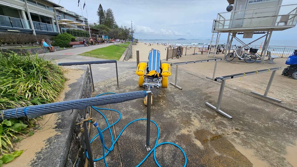 A concrete surface with sand scattered across it covers the foreground. In the middle is a striped blue-and-white beach wheelchair with yellow wheels, positioned between metal railings. On the left, there is vegetation near a stone wall, a paved pathway, and a grass lawn with people sitting on benches. On the right, a lifeguard tower is elevated on stilts, with bicycles and a blue bag underneath. In the background, a sandy beach leads to the water's edge, with people visible throughout.