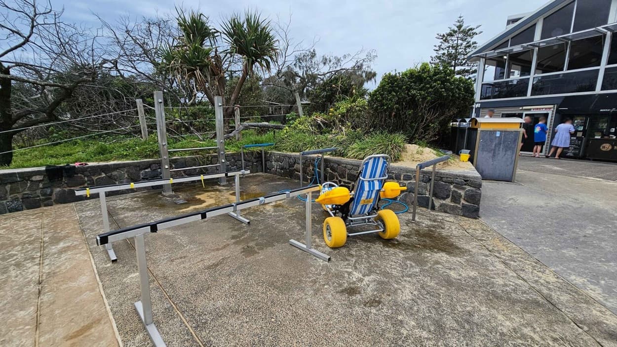 A wet concrete floor stretches across the foreground, with metal handrails forming a rectangular structure in the middle. On the right, a striped blue-and-white beach wheelchair with yellow wheels is placed near a dark stone wall. To the left, a grassy area with trees is bordered by another section of the stone wall. In the background, a multi-level glass-fronted building stands near a group of people and a yellow rubbish bin, with bushes and sand dunes nearby.