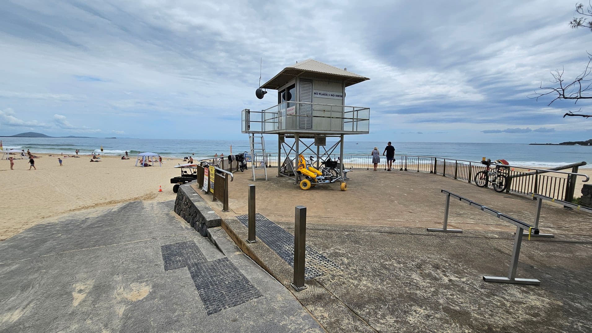 A concrete ramp with textured sections leads down to a sandy beach in the foreground. On the left, the beach extends with people, umbrellas, and small tents scattered across the sand. In the middle, a raised lifeguard tower with metal railings stands on a concrete platform, with a yellow beach wheelchair parked below. On the right, metal railings border the platform, alongside a bicycle and a small tree branch partially in view. The ocean stretches into the background.