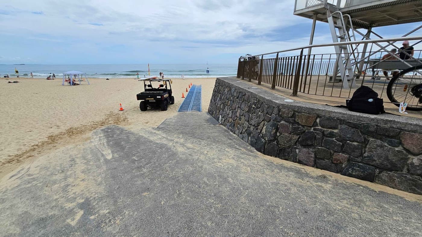 A concrete ramp in the foreground leads down to a sandy beach, with a textured mat extending across the sand in the centre. On the left, an orange traffic cone and a small utility vehicle are parked near the mat. On the right, a stone wall supports a raised platform with a metal railing, where a lifeguard tower and a bench are positioned. A black bag and part of a bicycle are visible next to the wall. In the background, people and beach items are scattered on the sand near the water.