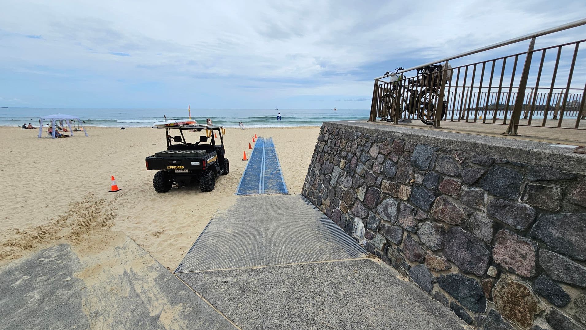 A concrete ramp in the foreground leads down to a sandy beach, with a textured mat extending across the sand in the centre. On the left, an orange traffic cone and a small utility vehicle are parked near the mat. On the right, a stone wall supports a raised platform with a metal railing, where a lifeguard tower and a bench are positioned. A black bag and part of a bicycle are visible next to the wall. In the background, people and beach items are scattered on the sand near the water.