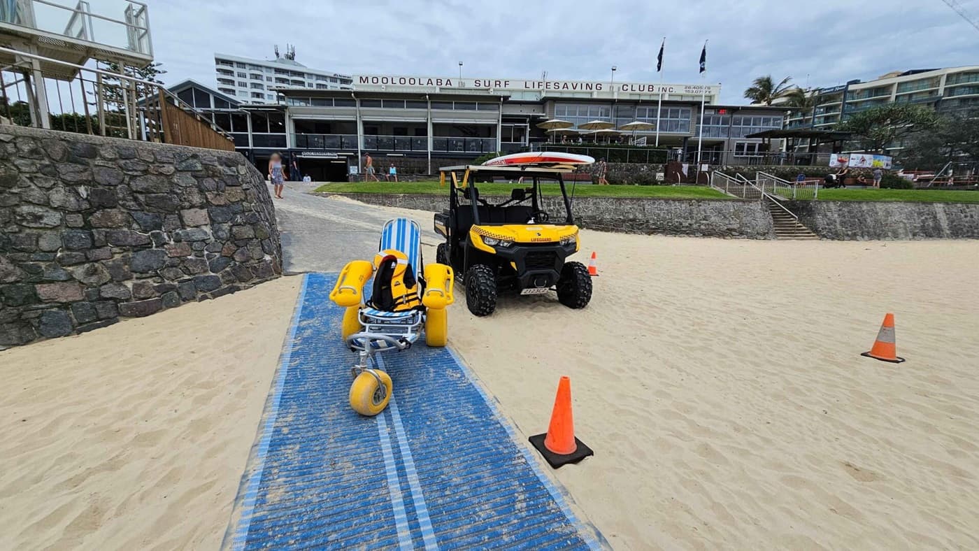 A blue textured mat stretches across the sandy beach in the foreground, with an orange traffic cone on either side. On the left, a stone wall curves upwards towards a wooden ramp. In the middle, a striped beach wheelchair with yellow wheels is parked beside a yellow all-terrain utility vehicle. On the right, additional cones are spaced on the sand. In the background, a grassed area leads to the Mooloolaba Surf Lifesaving Club building, with stairs and pathways bordered by stone walls.