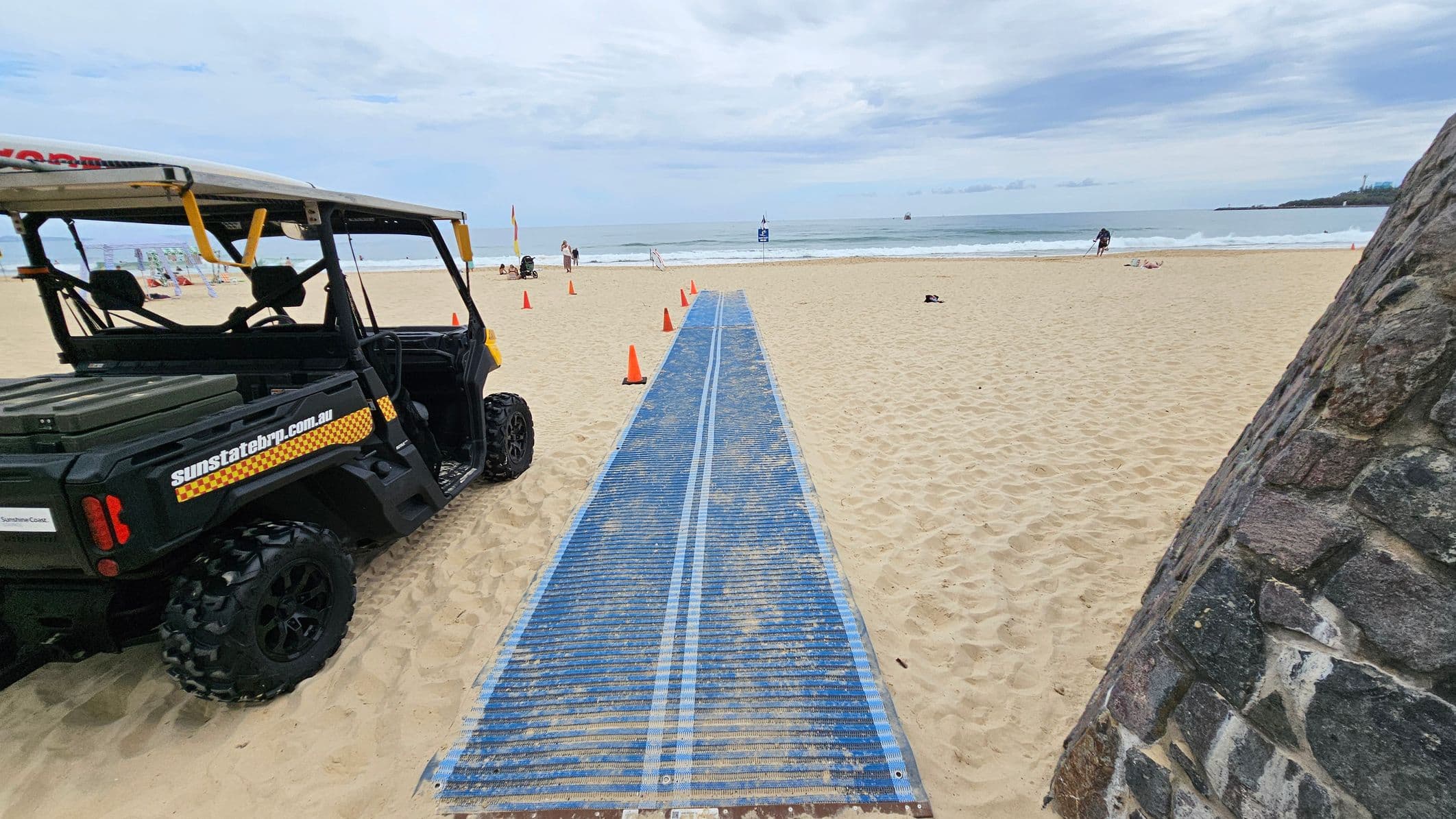 A blue textured mat stretches across a sandy beach from the foreground to the middle, flanked by orange traffic cones. On the left, a black off-road utility vehicle with "sunstatebrp.com.au" branding is parked on the sand. On the right, a rough stone wall extends into the foreground. In the background, the sand meets the shoreline with people standing near the water and signage visible close to the waves.