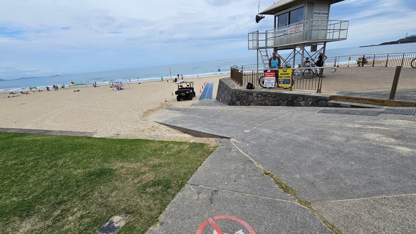 A concrete ramp with a no-entry sign leads down towards a sandy beach on the left, with grass in the foreground meeting the ramp. The beach has scattered people sitting, walking, and playing near the water. On the right, a raised lifeguard tower with railings stands on a stone base, surrounded by signs and bicycles. A paved pathway runs along the background near the tower, bordered by a black metal fence overlooking the beach.