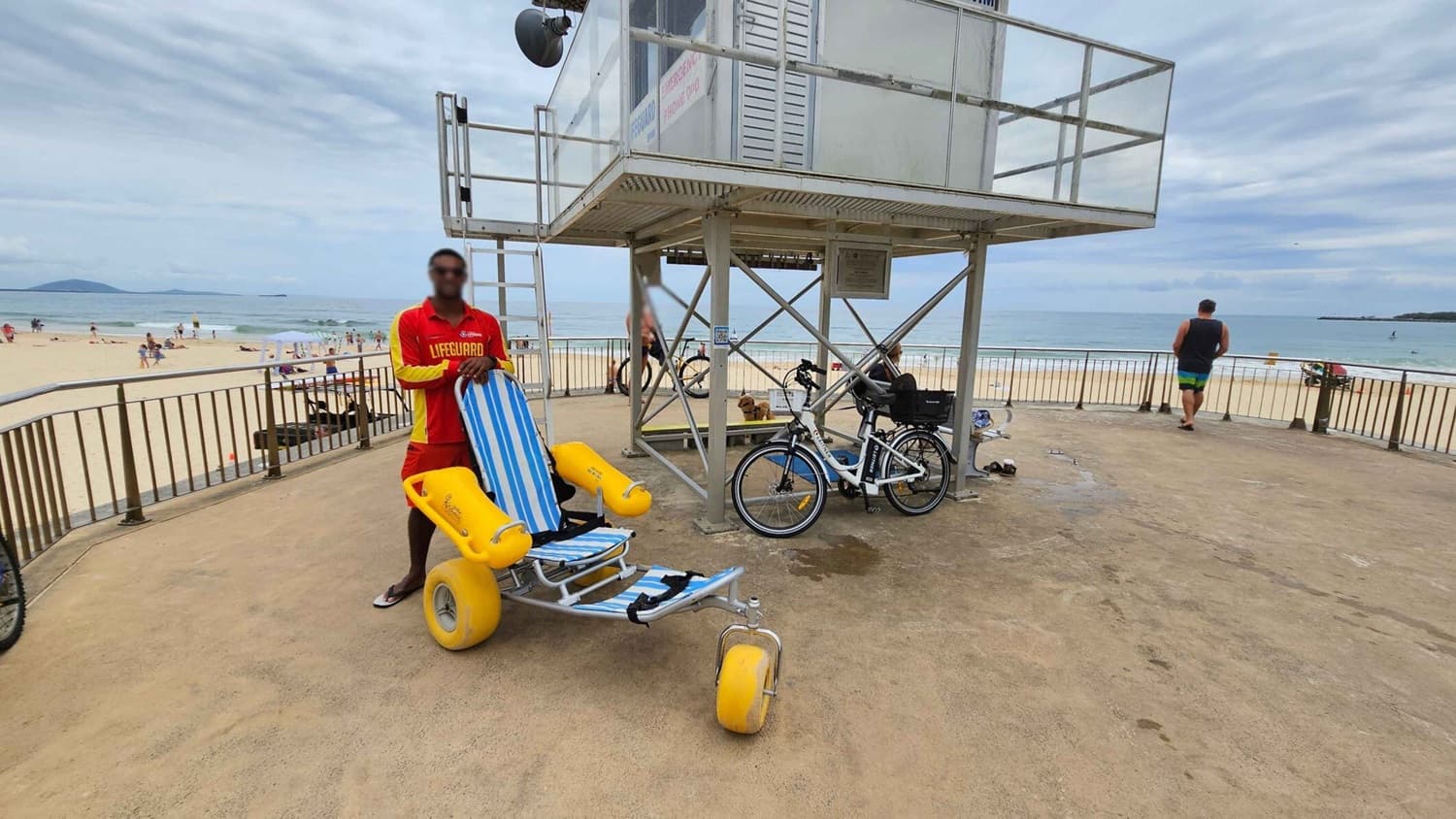 A lifeguard in a red and yellow uniform stands next to a beach wheelchair with large yellow wheels and a blue-striped seat. The lifeguard tower is elevated on metal supports, with a bicycle parked underneath. In the background, a sandy beach with people and the ocean is visible. A man in swimwear is walking towards the railing overlooking the beach. The ground is concrete, and the area is enclosed by a metal fence.