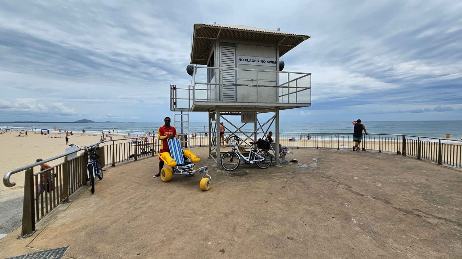 A concrete platform with a sandy texture has a lifeguard tower centred, raised on metal supports with a sign reading “No flags = No swim.” On the left, a lifeguard in a red shirt stands by a yellow beach wheelchair next to a parked bicycle, with railings overlooking the sandy beach. On the right, a man leans on the railings, facing the sea. In the background, people are scattered across the beach near the water.