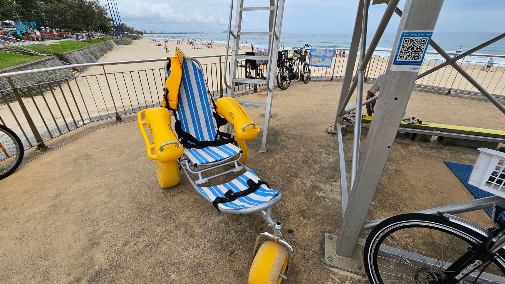 A striped blue and white beach wheelchair with yellow armrests and flotation wheels is positioned on a sandy concrete surface in the foreground, near a metal lifeguard tower on the right. To the left, a curved metal railing overlooks a sandy beach. In the middle, two bicycles lean against the railing. In the background, a grassy area slopes to a stone wall along the beach, with scattered beachgoers near the shoreline. A QR code sign is attached to the lifeguard tower.
