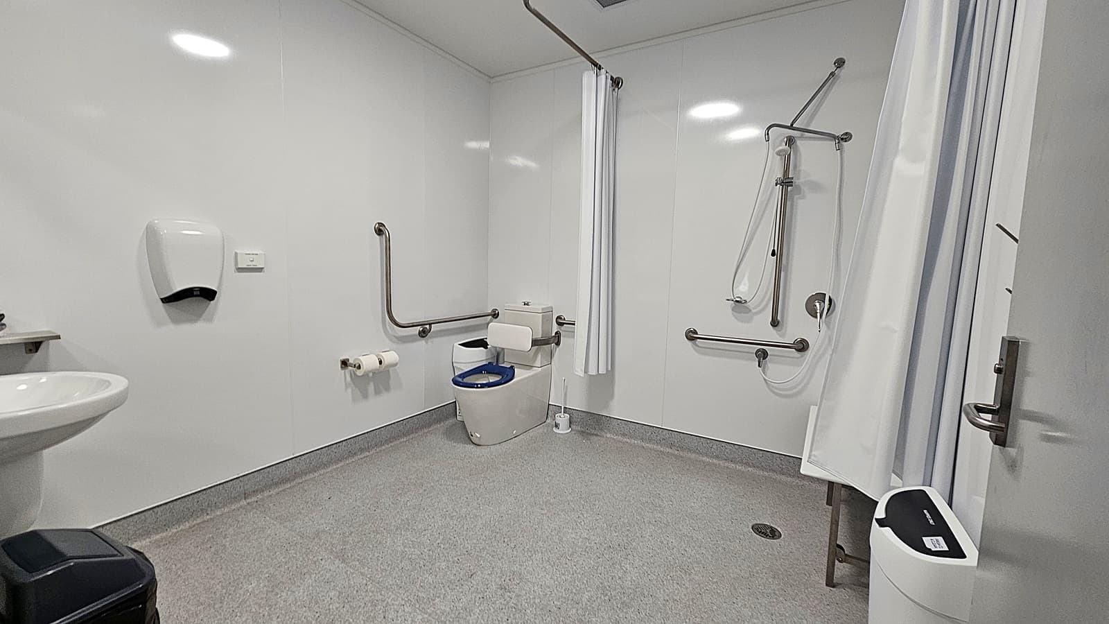 On the left, a white sink is mounted on the wall, with a black bin positioned beneath it. Above the sink, a soap dispenser is attached to the wall. The floor is a smooth grey surface, extending throughout the room. In the centre, a toilet with grab rails is visible, alongside a roll of toilet paper mounted on the wall. To the right, a shower area is enclosed by a curtain, with grab bars and a handheld showerhead.