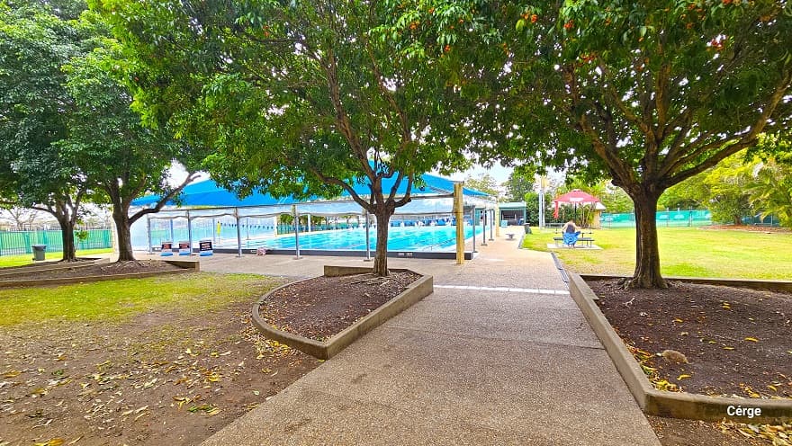 Step-free sloping pathway from the reception leads to the 25m, nine-lane lap pool at Murrumba Downs Swimming Pool. A large bright blue dome shade covers the closest section of the pool and has three signboards. A semi-circle and elongated triangular garden can be found around the area, which contains the trees that offer shade to the pathway. On the right side of the pool is a large field with a red sunshade with a table and bench.