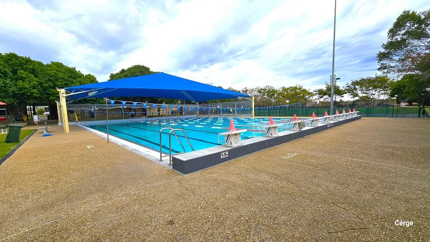 Starting point of the 25m, nine-lap pool at Murrumba Downs Swimming Pool. Each lane has a white diving block that is numbered from 1-9, and on top is an orange cone. Both the starting and end point of the lap pool has an elevated grey floor section. On each side of the pool are pool ladders marked by inverted U stainless steel handles. Strings of triangular blue flags overhang the pool.