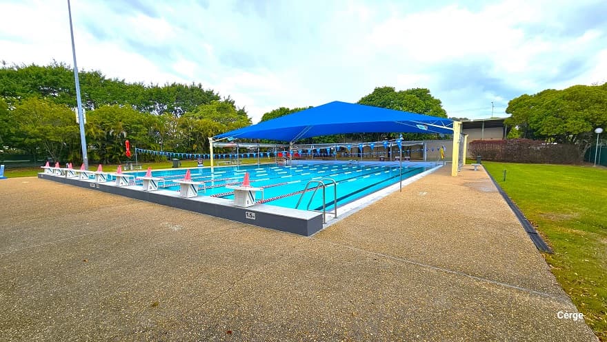 25m, nine-lane outdoor pool at Murrumba Downs Swimming Pool. The starting pool section has an elevated grey tile floor with nine white diving blocks that have orange traffic cones on top. The pool deck is brown with a rough surface to avoid slipping. The other end is partially covered by a wide blue sunshade supported by yellow metal frames. A few benches with tables are available around the pool, and strings of triangular blue flags are wrapped around steel posts on opposite pool sides.