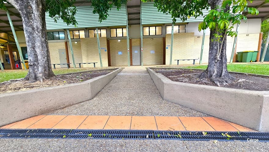 Ramp access to the accessible bathroom and changing rooms. The ramp access is between two triangular gardens in front of the bathrooms and is marked by a dark grey grating water channel and a single lane of light brown tile floor. The bathroom area has a corrugated lime green panel covering the ceiling. Grey benches are available in front. Towards the left end are large green garbage bins with black wheels.