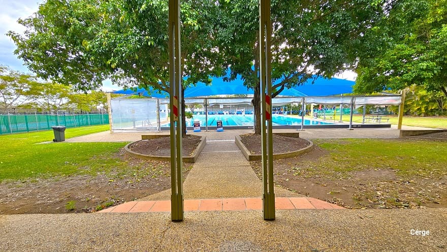 Pool granite pathway with a rough surface that is connected to the ramp access to the bathroom. Before reaching the pool, guests will pass through two semi-circular gardens with trees that have large canopy covers near the pool deck that has a large blue dome structure. There is a grey garbage bin on the left side of the dome covered by a white plastic cover. After the single lane of light brown tile floor are two grey posts with a narrow hole with orange and red stripe markings in the middle.