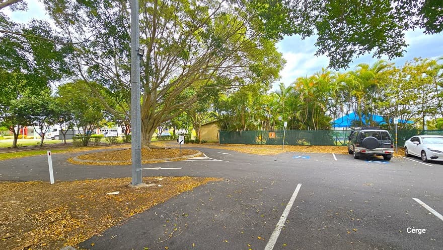 There are two accessible car parking spaces near the entrance to Murrumba Downs Swimming Pool which is close to the green hessian perimeter fence. It has a blue and white logo marking for persons with mobility disabilities. There is a circular garden with a large tree at the end of the car park. A white arrow can be seen on the left pointing inside, and another white arrow pointing outside and back to the carpark area. A road sign for ‘Keep Left’ can be seen in front of the circular garden.